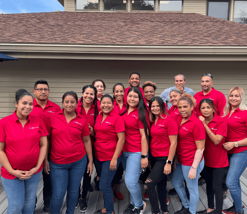 A group of 18 people wearing red shirts stands together on a deck, smiling at the camera, with a house in the background.